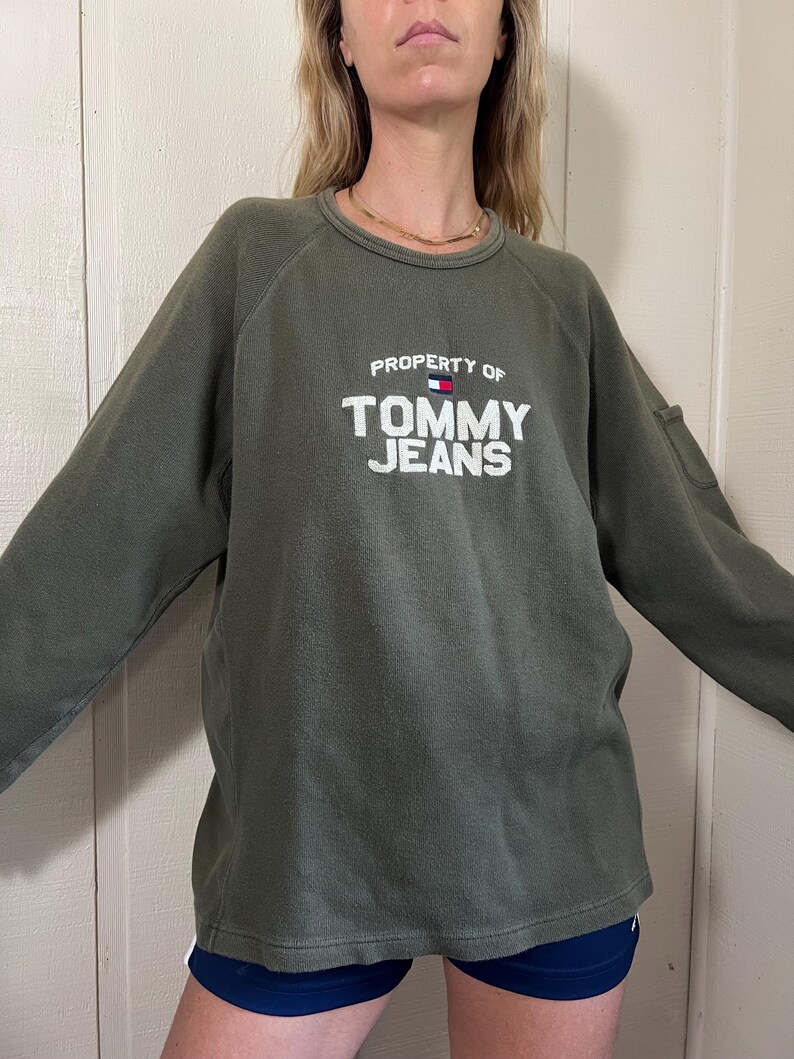 May include: Olive green sweatshirt with the text "PROPERTY OF TOMMY JEANS" in white block letters. The sweatshirt has long sleeves and a crew neck. A small flag graphic is above the text. The model is wearing navy blue shorts.