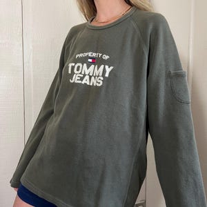 May include: Olive green sweatshirt with the text "PROPERTY OF TOMMY JEANS" in white. The sweatshirt has a small pocket on the sleeve and a crew neck. The model is wearing navy blue shorts.