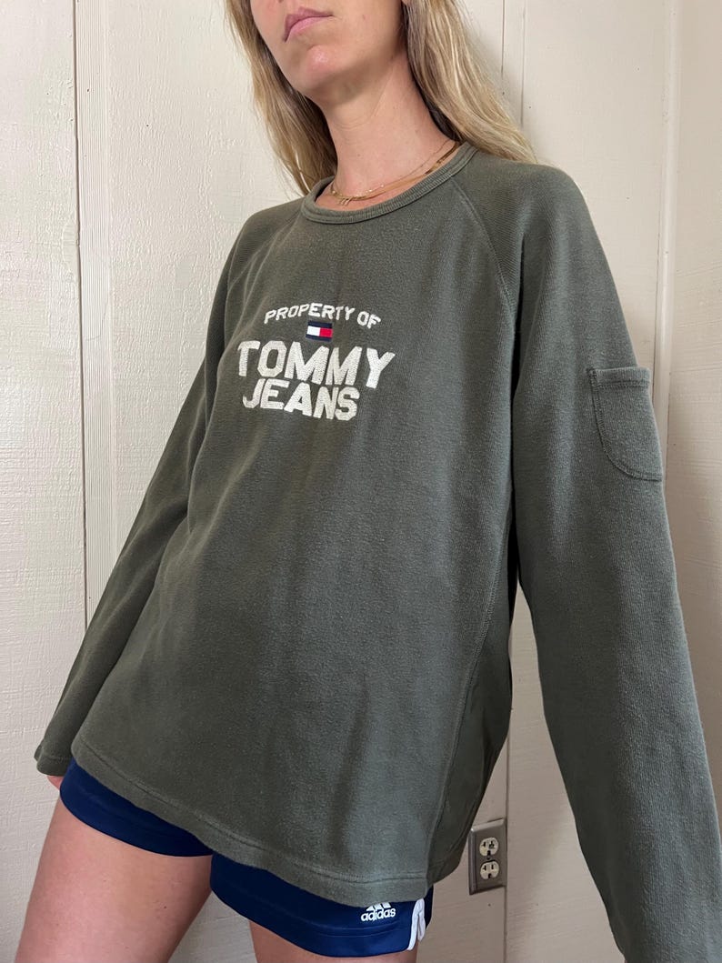May include: Olive green sweatshirt with the text "PROPERTY OF TOMMY JEANS" in white. The sweatshirt has a small pocket on the sleeve and a crew neck. The model is wearing navy blue shorts.