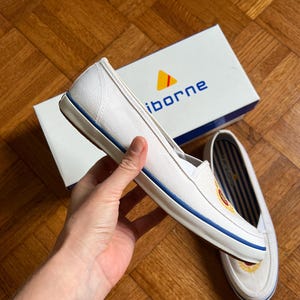 May include: White canvas slip-on shoes with a blue stripe along the sole and a logo on the side. The shoes are next to a white box with the brand name "iborne" printed on it.