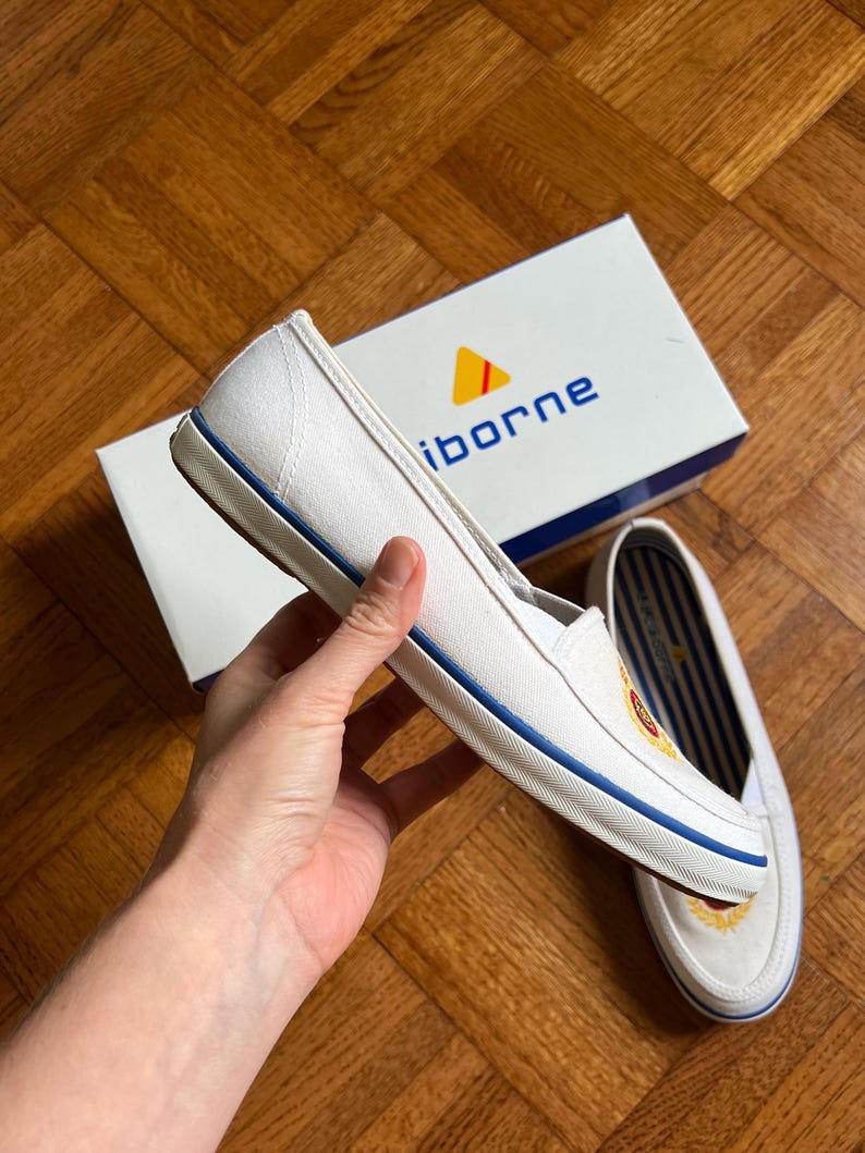 May include: White canvas slip-on shoes with a blue stripe along the sole and a logo on the side. The shoes are next to a white box with the brand name "iborne" printed on it.