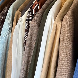 K&ouml;nnte beinhalten: Eine Sammlung verschiedener Jacken und M&auml;ntel, die an einer Stange h&auml;ngen. Die Kleidungsst&uuml;cke umfassen Denim-, Leder- und Tweed-Texturen in Braun-, Beige- und Gr&uuml;nt&ouml;nen. Einige M&auml;ntel haben Pelz- oder Shearling-Kragen.