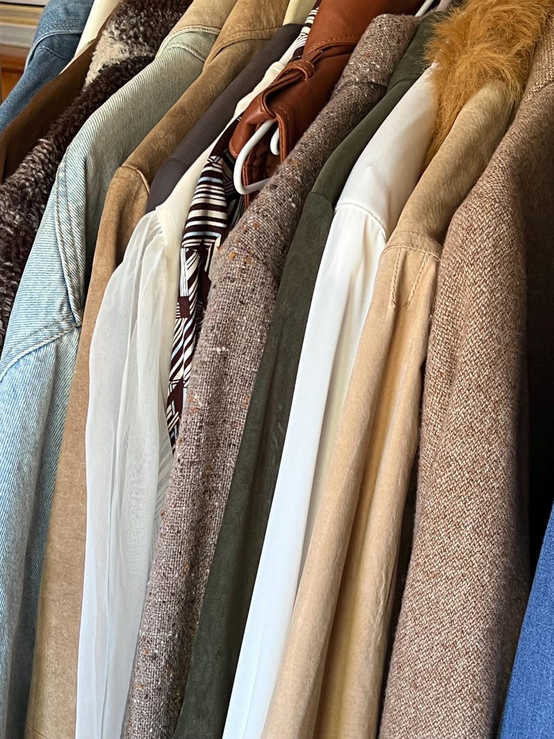 K&ouml;nnte beinhalten: Eine Sammlung verschiedener Jacken und M&auml;ntel, die an einer Stange h&auml;ngen. Die Kleidungsst&uuml;cke umfassen Denim-, Leder- und Tweed-Texturen in Braun-, Beige- und Gr&uuml;nt&ouml;nen. Einige M&auml;ntel haben Pelz- oder Shearling-Kragen.