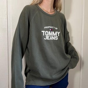 May include: Olive green sweatshirt with the text "PROPERTY OF TOMMY JEANS" in white block letters. The Tommy Hilfiger logo is above the word "JEANS". The sweatshirt has long sleeves and a crew neck.
