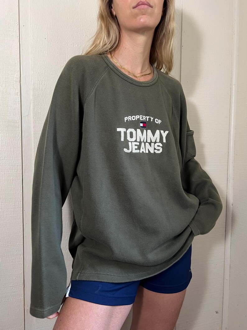 May include: Olive green sweatshirt with the text "PROPERTY OF TOMMY JEANS" in white block letters. The Tommy Hilfiger logo is above the word "JEANS". The sweatshirt has long sleeves and a crew neck.
