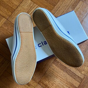 May include: Two white canvas plimsolls with blue edging and brown soles, presented on a wooden surface. The shoes are new and unworn, with a box partially visible in the background.