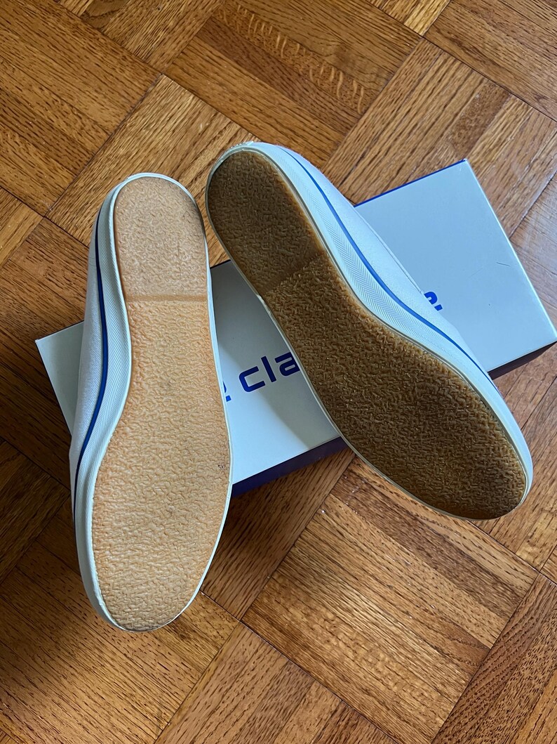 May include: Two white canvas plimsolls with blue edging and brown soles, presented on a wooden surface. The shoes are new and unworn, with a box partially visible in the background.