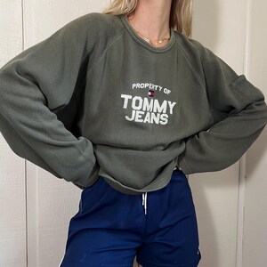 May include: An olive green sweatshirt with the text "PROPERTY OF TOMMY JEANS" in white. The sweatshirt is paired with navy blue athletic shorts with a white stripe down the side.