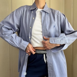 May include: A light blue long-sleeved button-down shirt with a small embroidered logo on the chest, worn over a white ribbed tank top and navy blue athletic shorts featuring a black and white striped elastic waistband with a white drawstring.