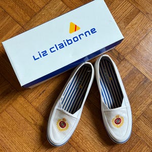 May include: White canvas slip-on shoes with blue and white striped interiors and a gold and red embroidered crest. The shoes are next to a white box with the brand name "Liz Claiborne" in blue lettering.