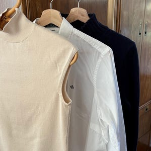 K&ouml;nnte beinhalten: Drei Kleidungsst&uuml;cke h&auml;ngen an Holzb&uuml;geln. Links ein &auml;rmelloser, hochgeschlossener beigefarbener Pullover. In der Mitte ein wei&szlig;es Button-Down-Hemd mit dem RL-Logo. Rechts ein marineblauer Pullover.