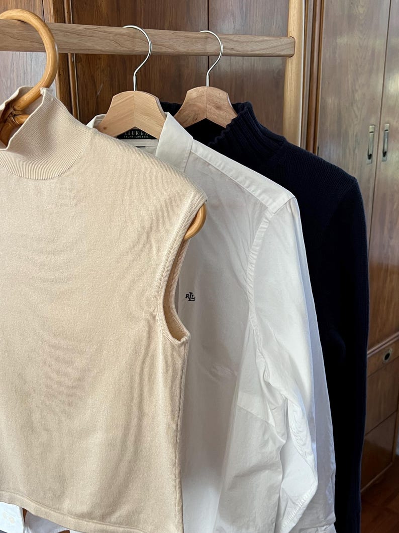 K&ouml;nnte beinhalten: Drei Kleidungsst&uuml;cke h&auml;ngen an Holzb&uuml;geln. Links ein &auml;rmelloser, hochgeschlossener beigefarbener Pullover. In der Mitte ein wei&szlig;es Button-Down-Hemd mit dem RL-Logo. Rechts ein marineblauer Pullover.