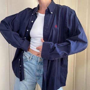 May include: A navy blue button-down shirt with a red logo on the chest. The long-sleeved shirt has white buttons and a classic collar. The shirt is worn open over a white ribbed tank top and light blue jeans.