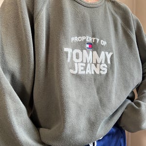 May include: Olive green sweatshirt with the text "PROPERTY OF TOMMY JEANS" in white. The sweatshirt has a crew neck and long sleeves. The Tommy Jeans logo is embroidered above the text.