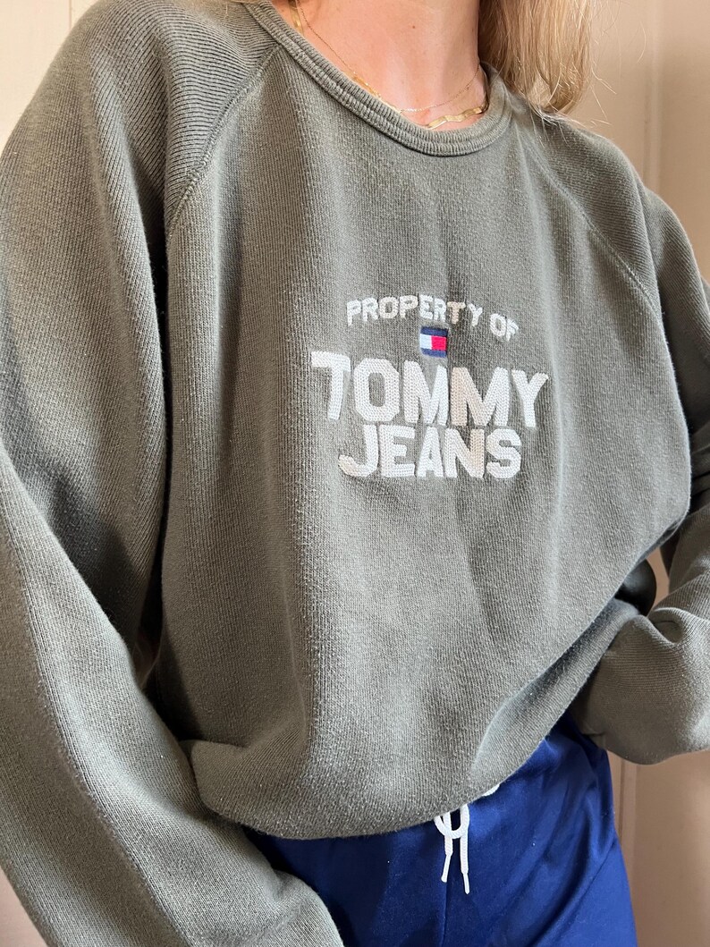 May include: Olive green sweatshirt with the text "PROPERTY OF TOMMY JEANS" in white. The sweatshirt has a crew neck and long sleeves. The Tommy Jeans logo is embroidered above the text.