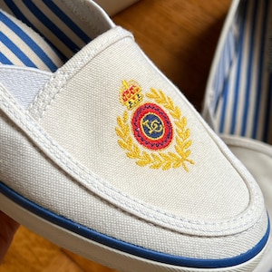 May include: White canvas slip-on shoes with blue trim and striped lining. The shoes have an embroidered crest in gold, red, and blue. The soles are white with a blue stripe.