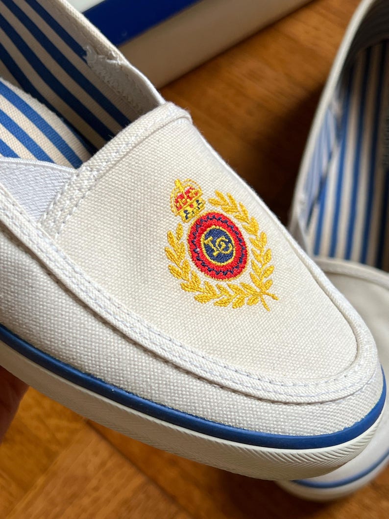 May include: White canvas slip-on shoes with blue trim and striped lining. The shoes have an embroidered crest in gold, red, and blue. The soles are white with a blue stripe.