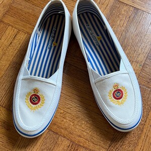 May include: A pair of white canvas slip-on shoes with blue trim and a gold and red crest emblem. The interior has blue and white striped fabric. The shoes are from the Liz Claiborne brand.