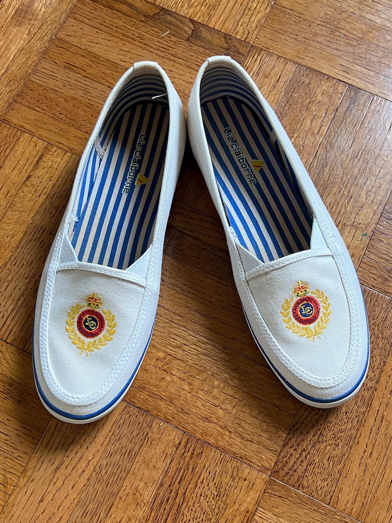 May include: A pair of white canvas slip-on shoes with blue trim and a gold and red crest emblem. The interior has blue and white striped fabric. The shoes are from the Liz Claiborne brand.