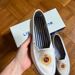 May include: White canvas slip-on shoes with a blue and gold embroidered crest. The shoes have blue and white striped insoles and a blue trim. The box in the background has the brand name "Liz Claiborne".
