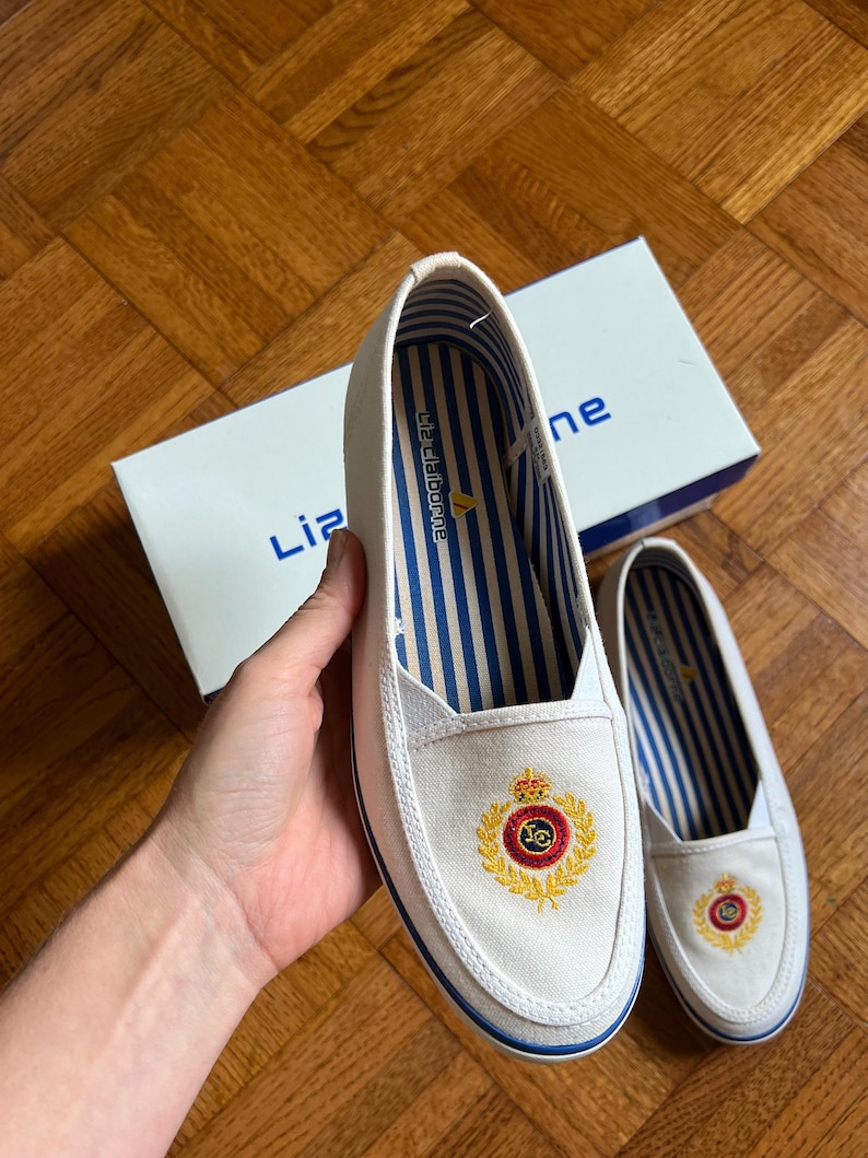 May include: White canvas slip-on shoes with a blue and gold embroidered crest. The shoes have blue and white striped insoles and a blue trim. The box in the background has the brand name "Liz Claiborne".