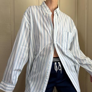 Könnte beinhalten: Ein langärmeliges Hemd mit Knopfleiste, weißem Grund und vertikalen hellblauen Streifen, klassischem Kragen und einer Brusttasche. Dazu wird eine dunkelmarineblaue Sportshorts mit weißem Kordelzug und elastischem Bund getragen.