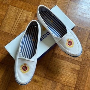 May include: White slip-on shoes with blue and white striped lining. Each shoe has an embroidered crest with red, gold, and black details. The shoes are on a white box with the brand name. The shoes are a classic design.