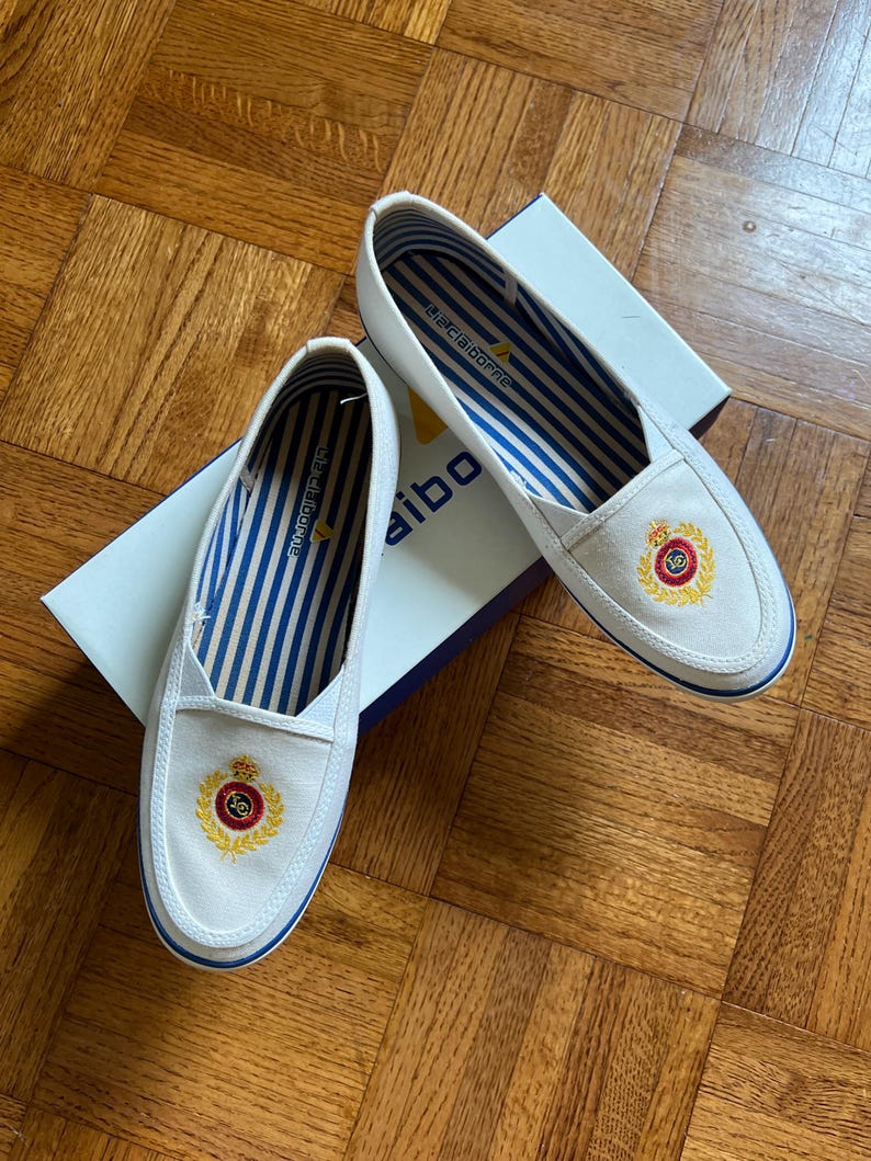 May include: White slip-on shoes with blue and white striped lining. Each shoe has an embroidered crest with red, gold, and black details. The shoes are on a white box with the brand name. The shoes are a classic design.
