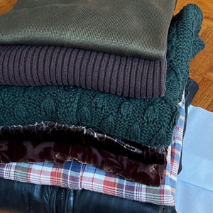 K&ouml;nnte beinhalten: Ein Stapel gefalteter Kleidungsst&uuml;cke in verschiedenen Farben und Texturen. Der Stapel umfasst einen dunkelgr&uuml;nen Pullover, einen gerippten lila Pullover, einen gr&uuml;n geflochtenen Pullover, einen gemusterten Schal, ein kariertes Hemd und eine schwarze Lederjacke.