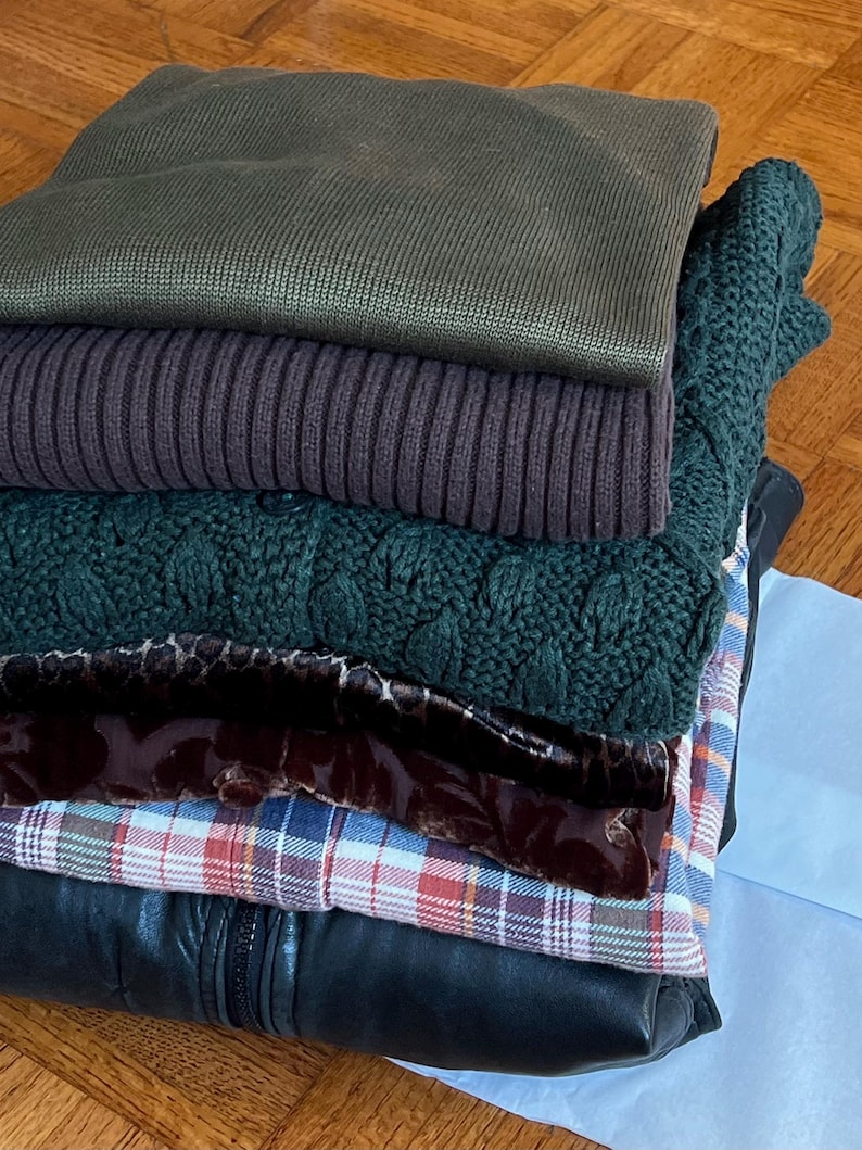 K&ouml;nnte beinhalten: Ein Stapel gefalteter Kleidungsst&uuml;cke in verschiedenen Farben und Texturen. Der Stapel umfasst einen dunkelgr&uuml;nen Pullover, einen gerippten lila Pullover, einen gr&uuml;n geflochtenen Pullover, einen gemusterten Schal, ein kariertes Hemd und eine schwarze Lederjacke.