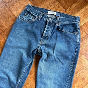 Pode incluir: Jeans bootcut de ganga azul com braguilha de botões e design clássico de cinco bolsos. Os jeans têm uma lavagem desbotada e a marca "GAP" é visível na cintura. Os jeans são feitos de ganga durável.