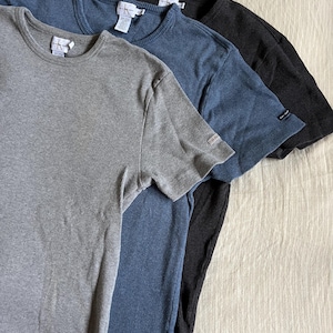 May include: Three short-sleeved ribbed knit shirts in gray, blue, and black. Each shirt has a crew neck and a small tag near the hem. The shirts are neatly arranged, showcasing their different colors and textures.
