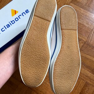 May include: Pair of white canvas trainers with blue detailing and brown rubber soles. The shoes are new and unworn, with a classic design. The brand name "claiborne" is visible on the box in the background.