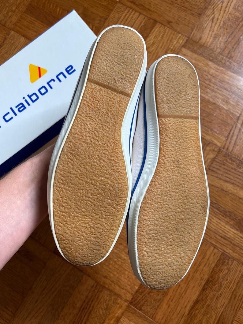 May include: Pair of white canvas trainers with blue detailing and brown rubber soles. The shoes are new and unworn, with a classic design. The brand name "claiborne" is visible on the box in the background.