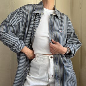 May include: A long-sleeve button-down shirt featuring vertical navy blue, yellow, and white stripes with a small Tommy Hilfiger logo on the chest pocket, worn over a plain white tank top and paired with white denim shorts with silver hardware.