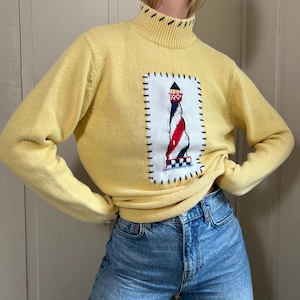 Può includere: Maglione giallo a collo alto con motivo faro. Il maglione presenta un quadrato bianco con un faro rosso, bianco e blu. Il faro è cucito sulla parte anteriore del maglione. Il maglione è abbinato a jeans blu.