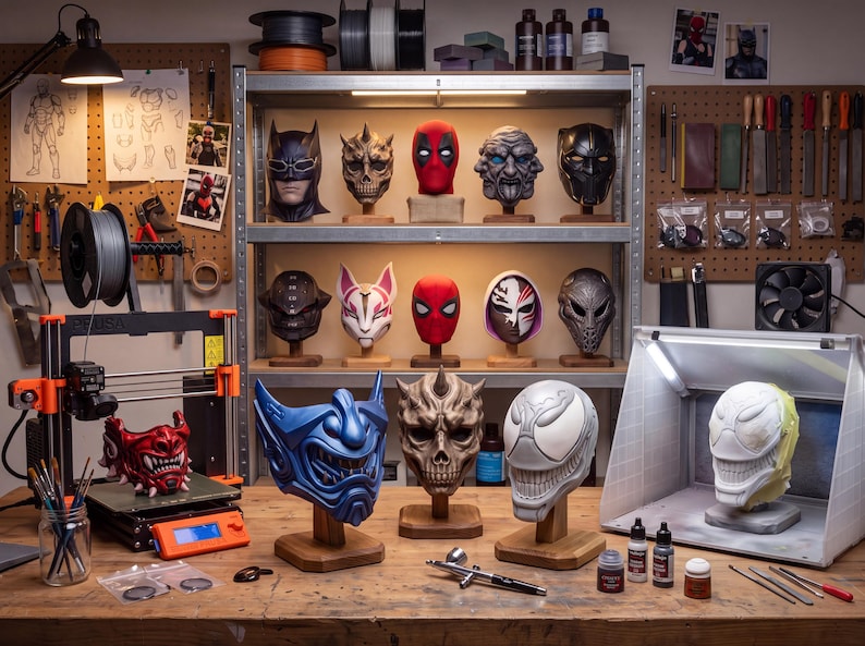 3D printing workshop with painted mask models, a desktop 3D printer, tools, paint supplies and finished masks displayed on shelves.
