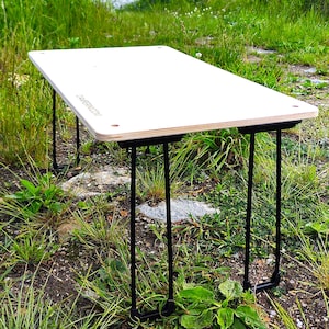 Può includere: Un tavolo rettangolare in legno chiaro con gambe in metallo nero. Il tavolo è allestito all'aperto su un letto di erba e piccole rocce. Il testo "CAMPERN.COM" è stampato sulla superficie del tavolo.
