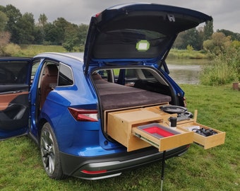 May include: A blue car with the back hatch open, revealing a wooden bed platform and drawers. The car is parked on a grassy area near a body of water.