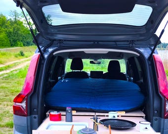 May include: A gray car with the back hatch open, revealing a blue inflatable mattress and a wooden platform with a portable stove, a kettle, and camping supplies.