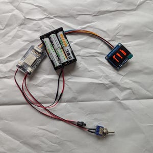 Puede incluir: Un circuito electr&oacute;nico con una peque&ntilde;a pantalla que muestra d&iacute;gitos rojos, conectado a un soporte de bater&iacute;a con dos pilas AAA. Una peque&ntilde;a placa de circuito y un interruptor tambi&eacute;n est&aacute;n conectados con cables. Los componentes est&aacute;n sobre una superficie blanca arrugada.