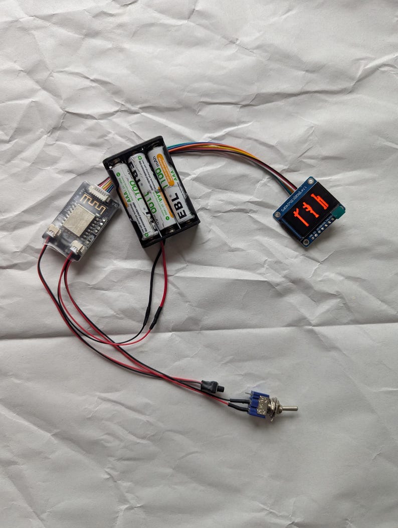 Puede incluir: Un circuito electr&oacute;nico con una peque&ntilde;a pantalla que muestra d&iacute;gitos rojos, conectado a un soporte de bater&iacute;a con dos pilas AAA. Una peque&ntilde;a placa de circuito y un interruptor tambi&eacute;n est&aacute;n conectados con cables. Los componentes est&aacute;n sobre una superficie blanca arrugada.