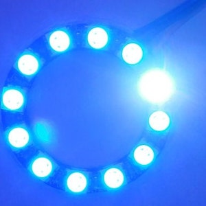 Puede incluir: Una tira de luces LED circular con luces blancas brillantes encerradas en un material azul flexible. Las luces están espaciadas uniformemente alrededor del círculo, emitiendo un brillo azul brillante. El fondo es azul liso.