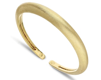 Puede incluir: Un brazalete de puño dorado con un acabado mate. El diseño abierto presenta una parte superior redondeada y un interior liso. El brazalete tiene aproximadamente 6,35 cm de diámetro.