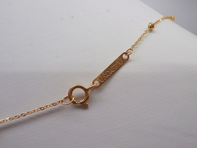 May include: Close-up of a delicate gold necklace with a small chain and a round clasp. The necklace features a small rectangular tag with the word "UNOAErre" engraved on it. The necklace is on a white background.