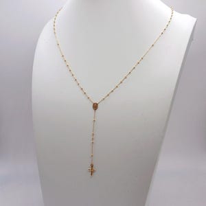 May include: A gold-colored rosary necklace displayed on a white mannequin. The necklace features small beads, a central oval pendant, and a cross pendant at the end. The chain is delicate and hangs in a Y-shape.