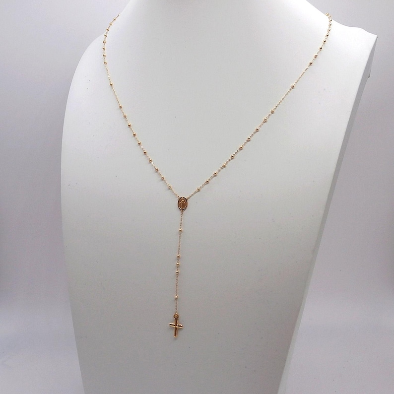 May include: A gold-colored rosary necklace displayed on a white mannequin. The necklace features small beads, a central oval pendant, and a cross pendant at the end. The chain is delicate and hangs in a Y-shape.