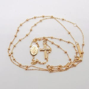 May include: Gold-colored rosary necklace with a cross pendant, a medallion, and small round beads. The necklace is displayed on a white surface. The cross and medallion are detailed with religious imagery.