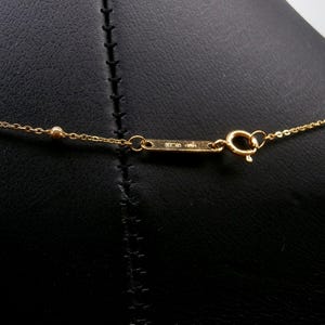 May include: Close-up of a gold necklace clasp and chain. The delicate chain features small, round beads. The clasp is rectangular with engraved markings. The background is a black surface.