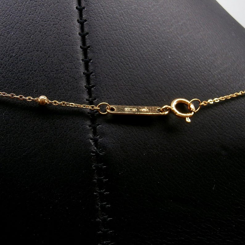 May include: Close-up of a gold necklace clasp and chain. The delicate chain features small, round beads. The clasp is rectangular with engraved markings. The background is a black surface.